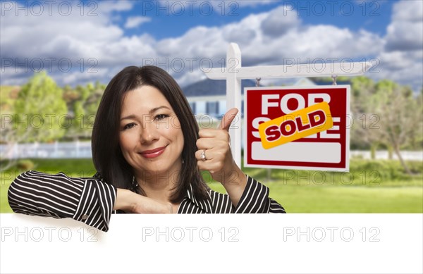 Pretty hispanic woman leaning on white with thumbs up in front of beautiful house and sold for sale real estate sign