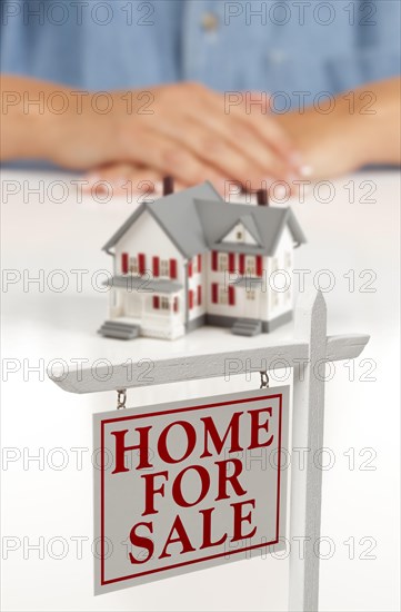 Womans folded hands behind model house and home for sale real estate sign in front on white surface