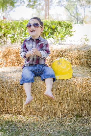 Cute young mixed-race boy laughing with sunglasses and hard hat outside sitting on hay bale