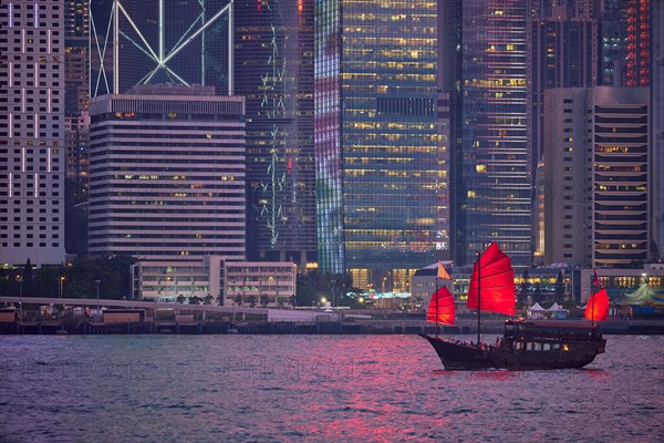 Tourist junk boat ferry with red sails and Hong Kong skyline cityscape downtown skyscrapers over Victoria Harbour in the evening. Hong Kong