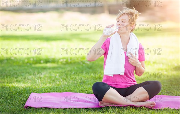 Young fit adult woman outdoors on her yoga mat with towel drinking from her water bottle