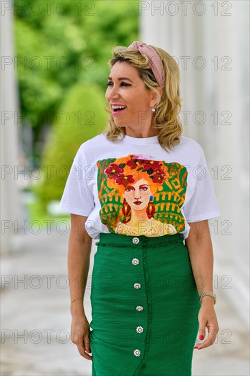 Portrait in einem Park einer huebschen Dame in ihren vierziger Jahren mit langen blonden Haaren. Sie geht auf die Kamera zu und blickt mit ueberraschte Blick zu Seite. Ein gruener langer Rock mit bedr...