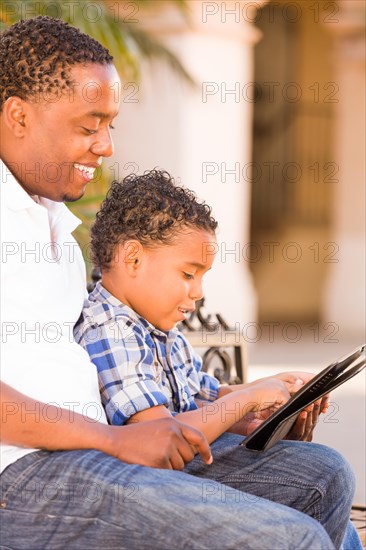African american father and mixed-race son using computer tablet on bench in park