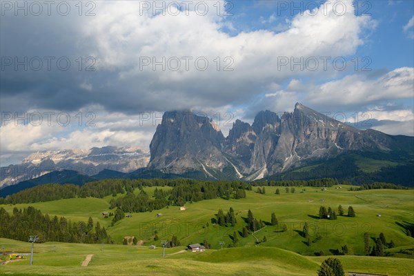 In Suedtirol auf der Seiser Alm im Fruehsommer mit einigen Almhuetten und einem Schilift auf saftig gruenen Weiden. Im Hintergrund ist ein grosser felsiger Berg mit einigen Wolken am blauem Himmel