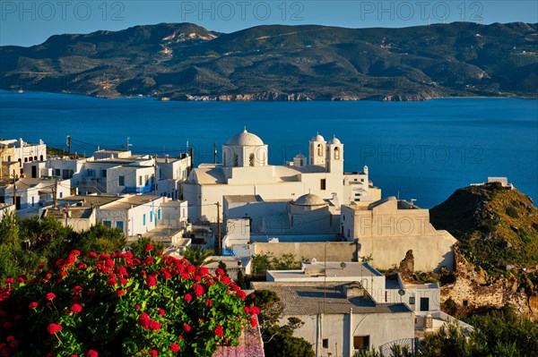Picturesque scenic view of Greek town Plaka on Milos island over red geranium flowers and Orthodox greek church