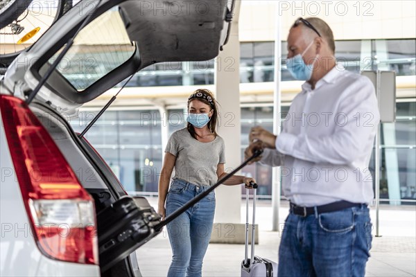 A taxi or Uber driver helping a passenger in a protective mask with her luggage at the airport