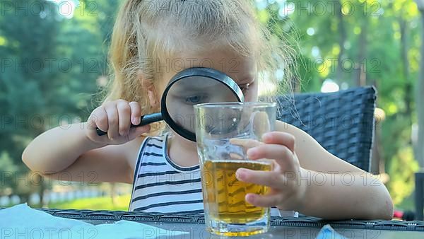 Cute little girl carefully looks into the lens at the juice in the glass. Close-up of blonde girl studying her juice in glass while looking at her through magnifying glass while sitting in street cafe...