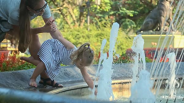 Mom holds smiling little daughter playing with splashes of water in the fountain. Mom with little girl play in fountain in the park and smiles. Odessa Ukraine