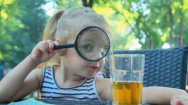 Little girl looks into the lens around. Close-up of blonde girl studying the world around her looking at it through magnifying glass while sitting in street cafe on the park. Odessa Ukraine