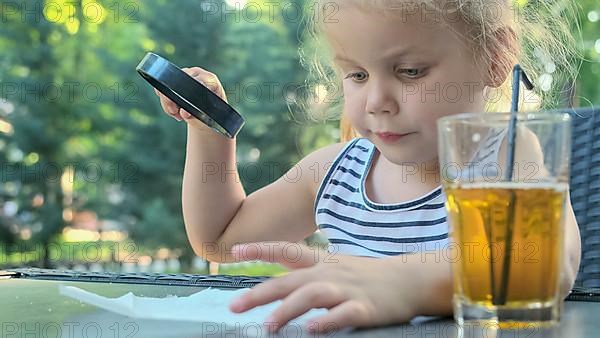 Little girl carefully looks into the lens at the salt. Close-up of blonde girl is studying salt crystals while looking at her through magnifying glass while sitting in street cafe in the park. Â Odess...