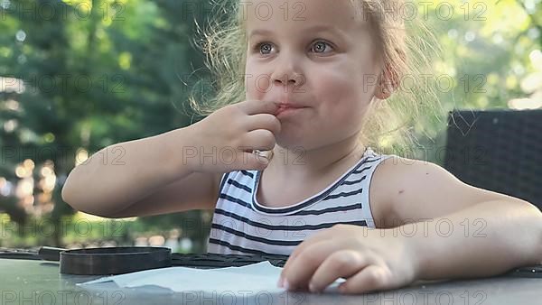 Little girl tastes salt. Close-up portrait of blonde girl takes salt from napkin with her finger and tastes it while sitting in street cafe on the park. Odessa Ukraine