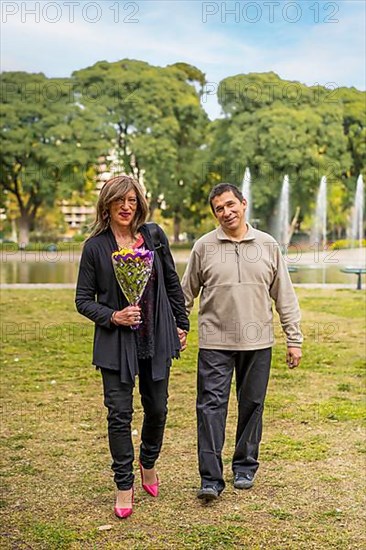 Mature transgender woman walking hand in hand with her boyfriend in a park happy because he gave her flowers,