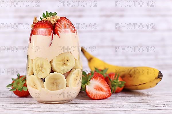 Healthy banana and strawberry fruit summer dessert with yogurt and granola layered in glass surrounded by fresh ingredients,