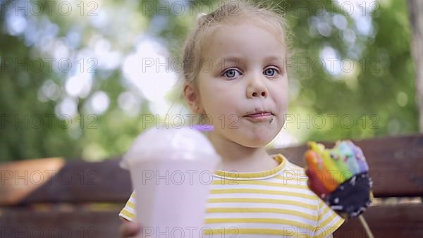 Ittle girl eats a colorful gingerbread and holds a milkshake in her hand. Close-up of cute child girl sitting on park bench and eating cookies with a milkshake and looks into the camera lens. Odessa, ...
