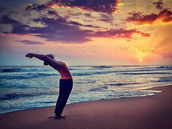 Vintage retro effect hipster style image of young sporty fit woman doing yoga Sun salutation Surya Namaskar pose Hasta Uttanasana on tropical beach on sunset