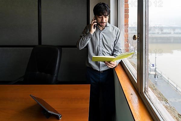 Latino businessman working in an office overlooking the river. Talking on the phone and reviewing notes