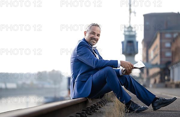 Man in blue suit sitting on railway track reading newspaper