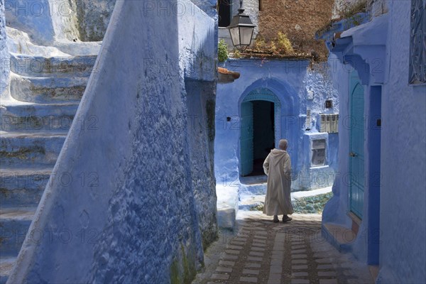 Man with djellaba walking next to blue buildings and staircase in an alley of the city