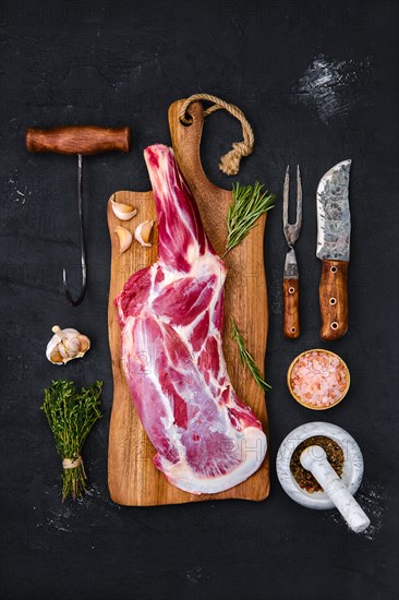 Fresh raw lamb leg bone in on wooden cutting board with herbs and seasoning