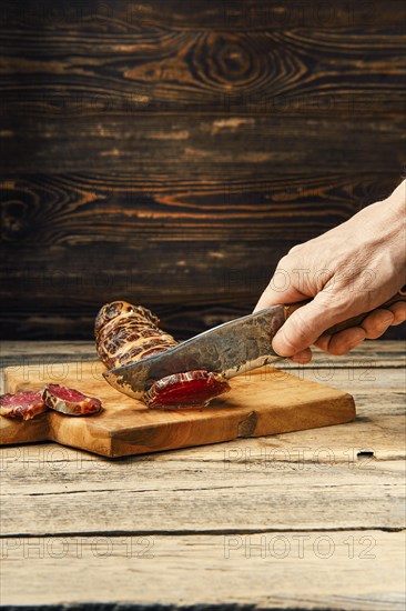 Hand with knife cutting smoked dried beef meat on slices on wooden cutting board
