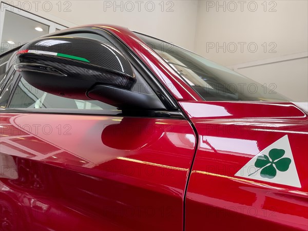 Detail of rear view mirror with carbon optic fairing with coloured stripes of Italian national colour flag Tricolore historical modern signet logo marking of Alfa Romeo Quadrifoglio green four-leaf cl...