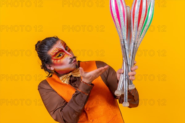 Kiss and portrait of happy juggler man in makeup vest juggling clubs on yellow background