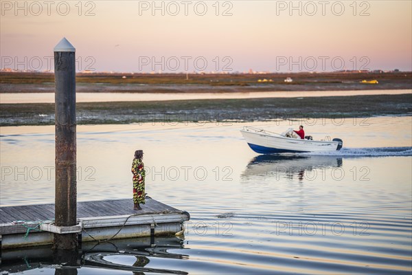A woman on a pier looking toward a man on the boat, early morning in Faro, Algarve