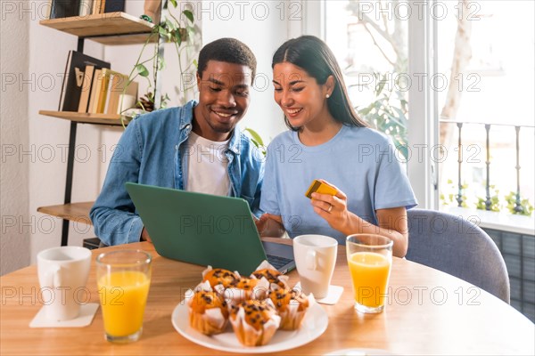 Couple making purchase online with the computer while having breakfast, next to the window, shopping on the internet