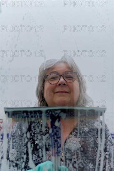 Close-up of a white-haired woman cleaning soap-covered windows with a squeegee