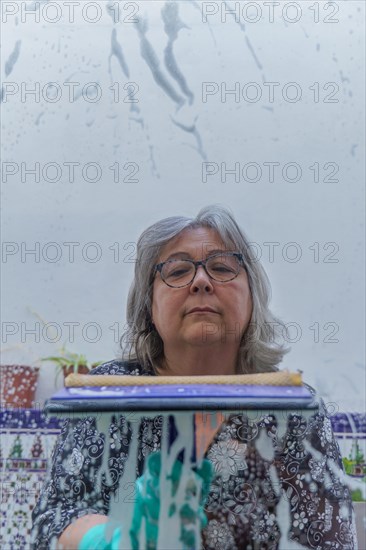 Close-up of a white-haired woman cleaning soap-covered windows with a squeegee
