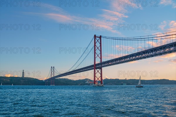 View of 25 de Abril Bridge famous tourist landmark of Lisbon connecting Lisboa to Almada on Setubal Peninsula over Tagus river with tourist yacht boats passing under. Lisbon