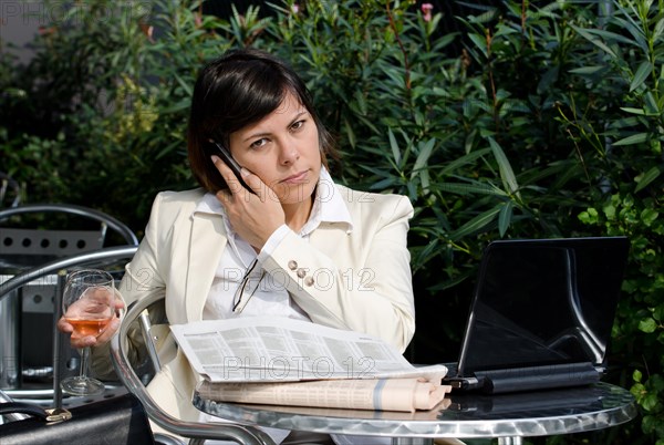 Business Woman Sitting with Her Personal Computer and Financial Newspaper and using Smart Phone on a Table Outdoor