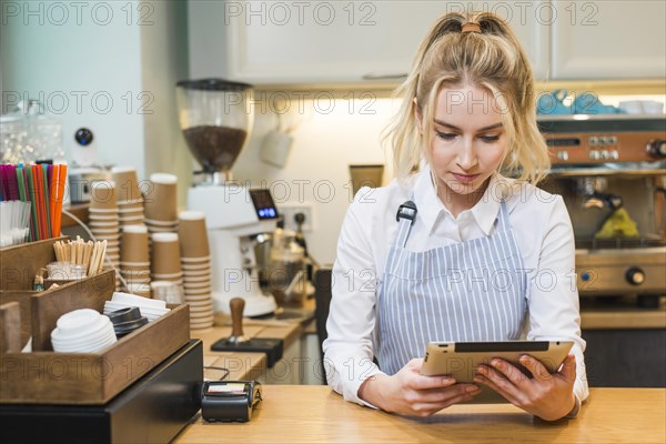 Blonde young woman standing coffee shop counter looking digital tablet