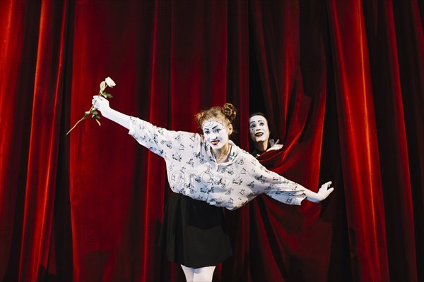 Female mime holding white rose performing stage with male mime peeking curtain