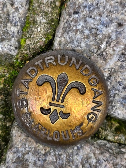 Brass-coloured metal medal embedded in a cobblestone street. The medal is circular and has a fleur-de-lis symbol in the centre. The medal is inscribed with the words Stadtrundgang and Saarlouis around...