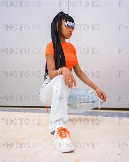A young trap dancer with braids. Black ethnic girl with orange t-shirt and cowboy pants on a gray background