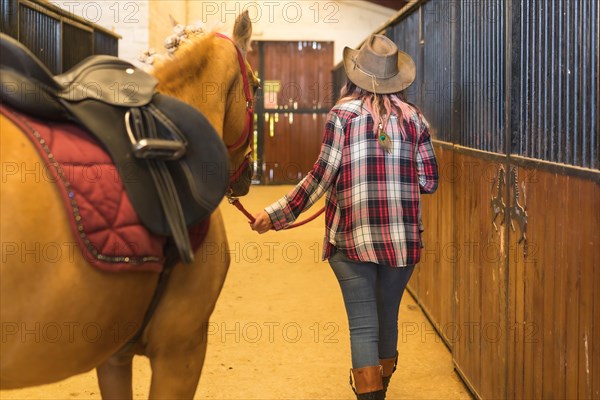 Unrecognizable cowgirl woman strolling with a horse in a stable