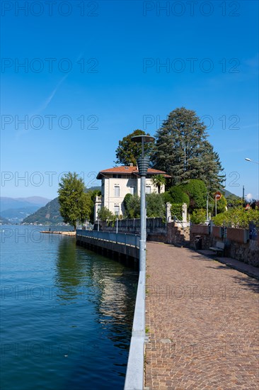 Walkway with Lamps and Railing and a House on the Waterfront on Lake Lugano with Mountain and Trees in a Sunny Summer Day in Porto Ceresio