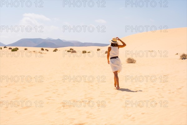 A young Caucasian tourist in a white dress and a hat walking through the dunes of the Corralejo Natural Park