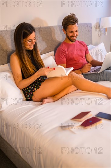 A handsome Caucasian couple lounging on the hotel bed and looking at a computer and a book. Summer lifestyle