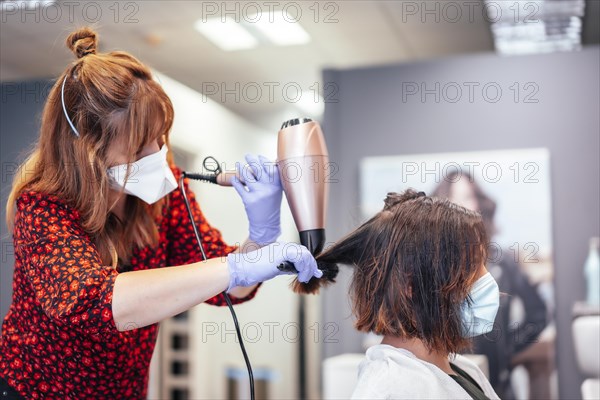 Blonde hairdresser with mask and gloves straightening client's brown hair with blow dryer. Reopening with security measures for hairdressers in the Covid-19 pandemic