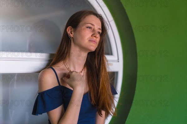 A young pretty redhead Caucasian girl smiling sitting in a blue dress next to a white sale of a green house