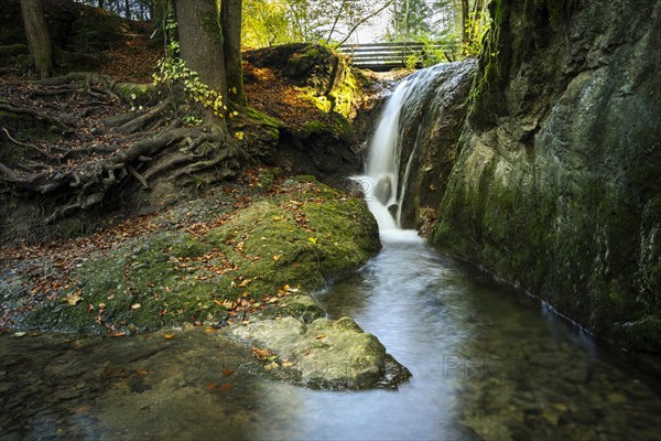 A small waterfall near the Gerats waterfall with moss-covered rocks and tree roots. The sun is shining in the background. Gerats