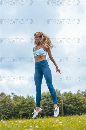 Fitness session with a young blonde caucasian woman exercising in nature with a blue maya on her feet and a white short shirt. Squatting and jumping