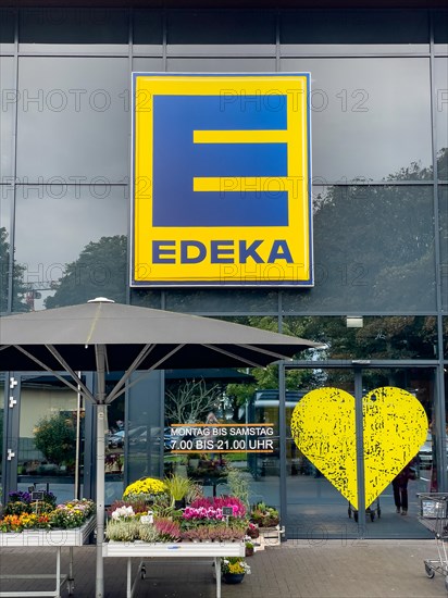 Logo with letter and lettering above entrance to supermarket below note on opening hours Monday to Saturday 7.00 a.m. to 9.00 p.m. to the right stylised heart of retail chain supermarket chain Edeka f...