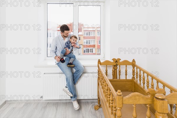 Father carrying his baby boy son sitting window sill looking wooden empty crib