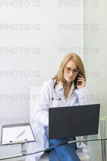 Female doctor talking on the phone to a patient while looking at her medical records on laptop in medical office