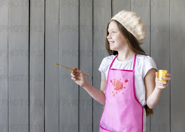 Side view smiling girl holding yellow paint bottle paintbrush hand standing against grey wooden wall