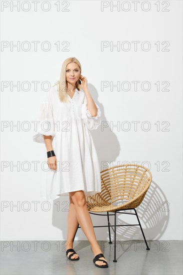 Beautiful woman in white sundress standing by wicker chair in sunny room