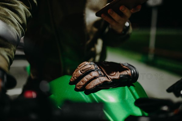 A brown leather motorcyclist glove lies on the tank of a green motorcycle while the motorcyclist looks at the travel route